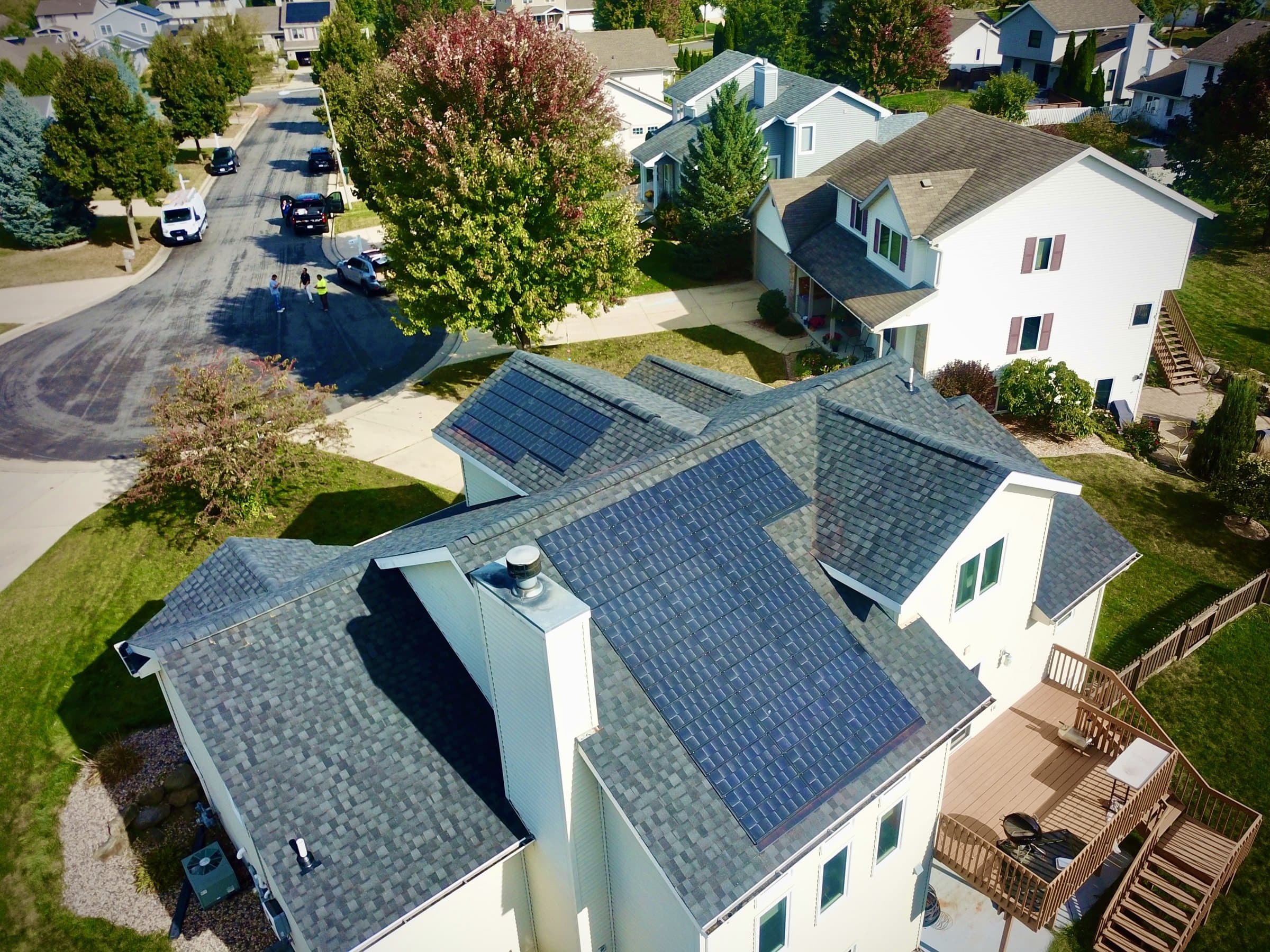 Aerial view of a detached house with a dark solar roof, surrounded by trees on a residential street.