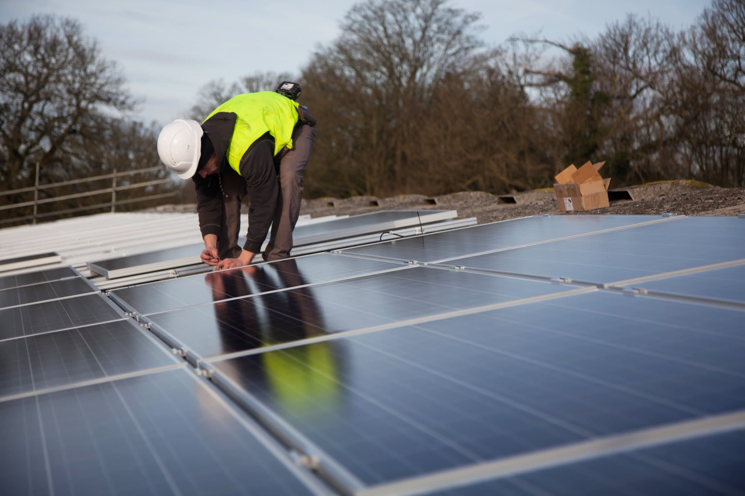 An installer in a hard hat and high-visibility jacket fitting a solar panel on a roof.